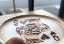 Classic vs. Modern: Which Card Games Are Winning in 2025? Close-up of a latte with Queen of Hearts art, a thumb in the foreground.